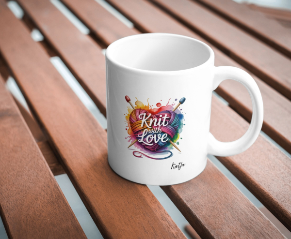 Weiße Tasse mit buntem Wollherz und dem Spruch „Knit with Love“, auf Holztisch fotografiert.